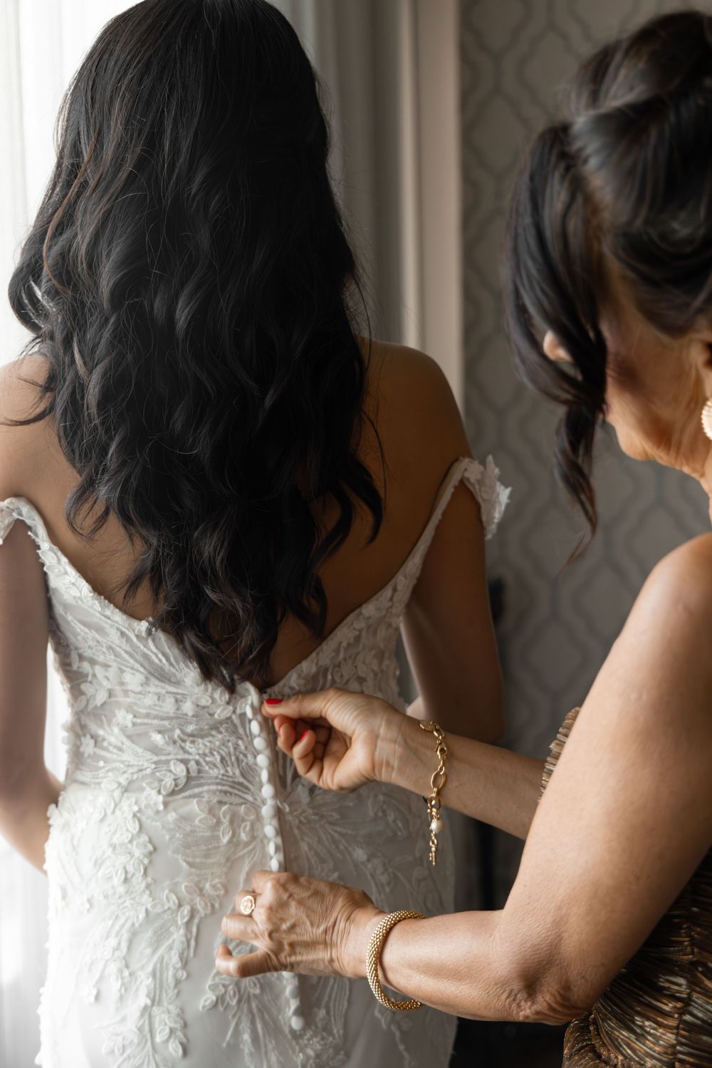 Someone helps zip up a white lace wedding dress with delicate details and buttons down the back.