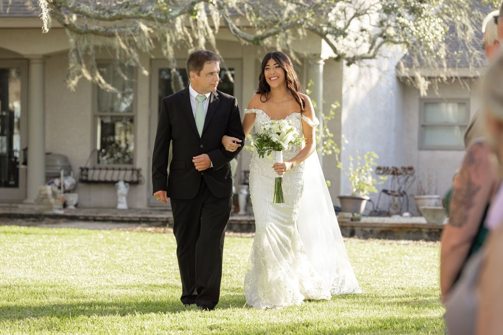 A wedding ceremony procession walks across a sunlit lawn between white trees at an elegant outdoor venue.