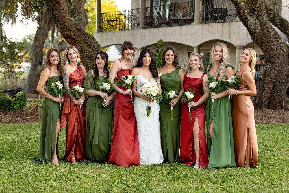 Wedding party wearing alternating red and green dresses poses together outdoors under trees.