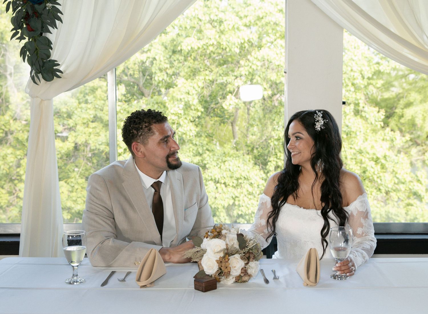A wedding party sits at a long white-draped table under an elegant outdoor pavilion during a reception celebration.