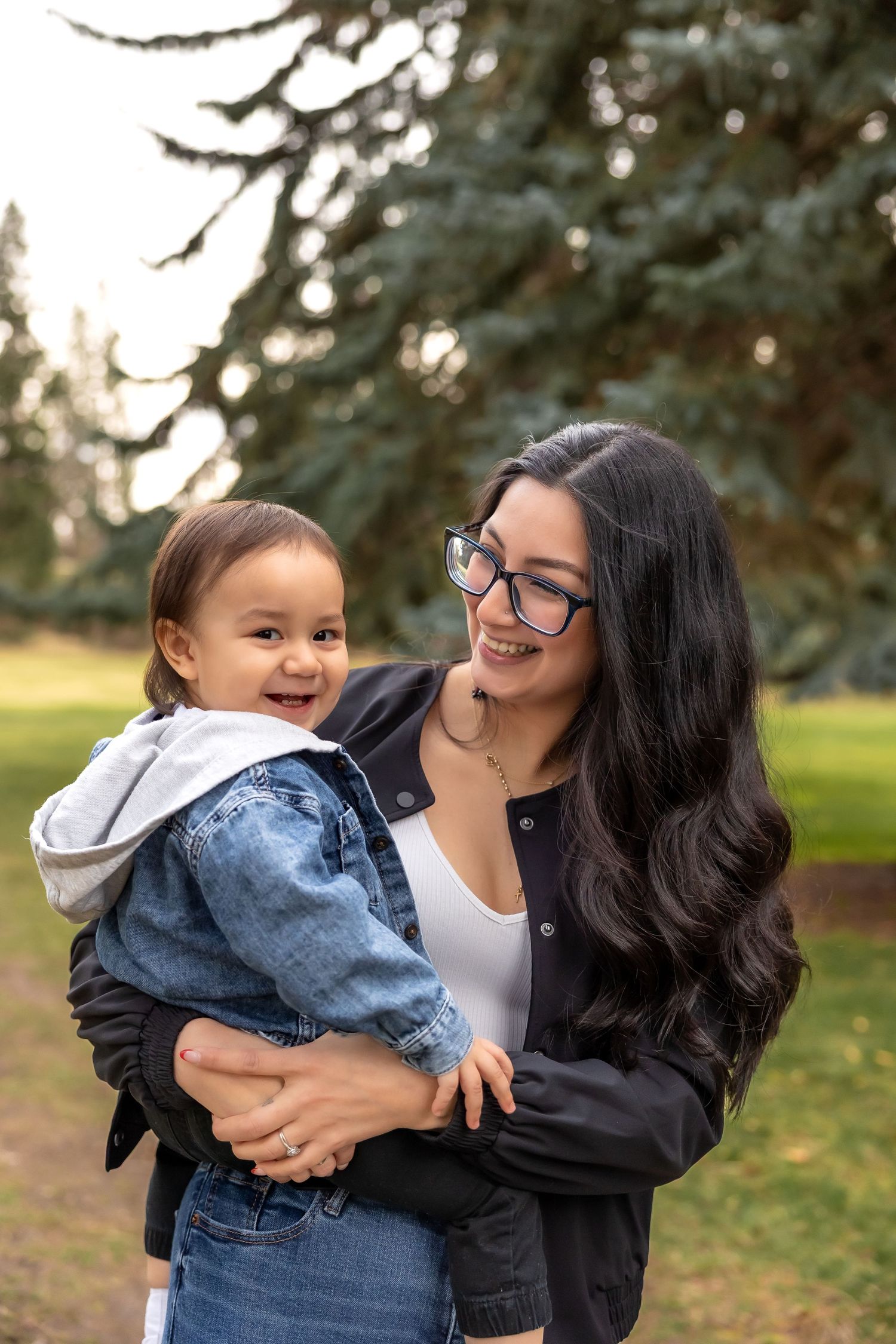 A joyful outdoor portrait of a parent and toddler sharing a laugh together in a park with evergreen trees.