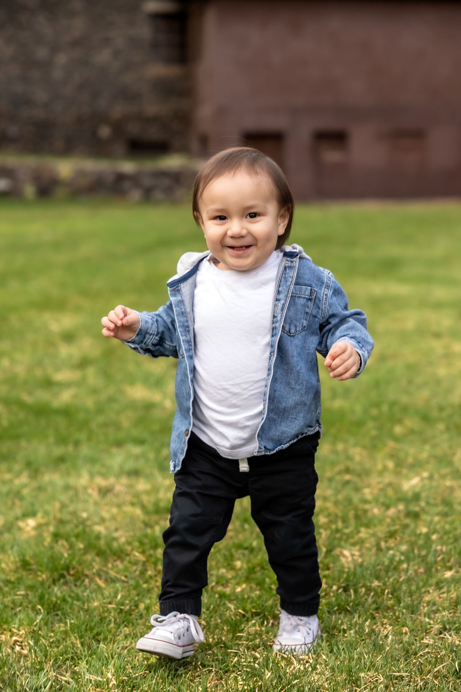 Small child in denim jacket and dark pants stands smiling on grass while learning to walk outdoors.