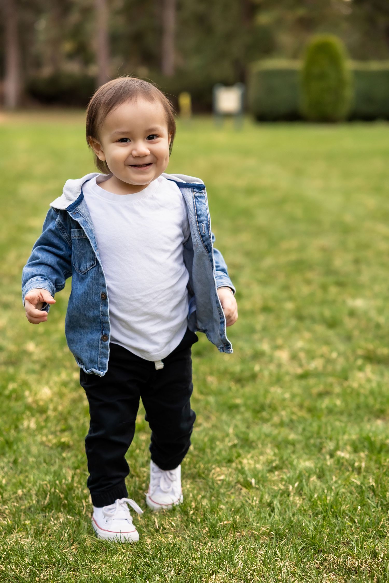 A small child in a denim jacket and white sneakers runs joyfully across a grassy lawn on a sunny day.