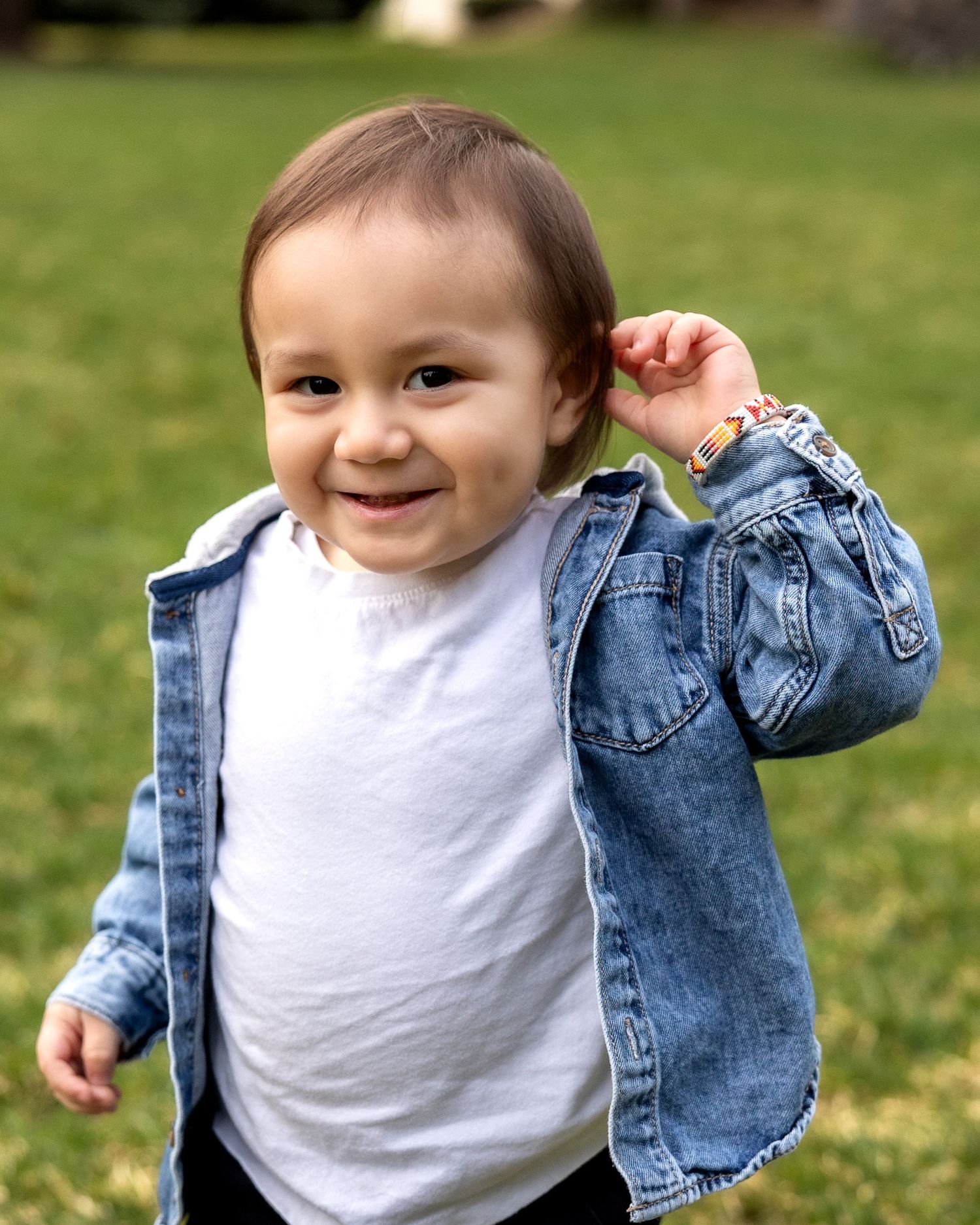 Small child wearing white t-shirt and denim jacket plays outdoors on green grass with joyful expression.