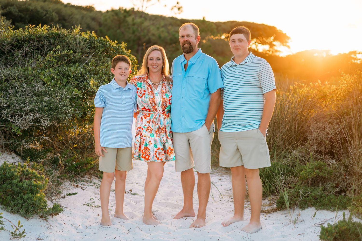 Family of four standing together at golden sunset among lush greenery on a sandy beach path.
