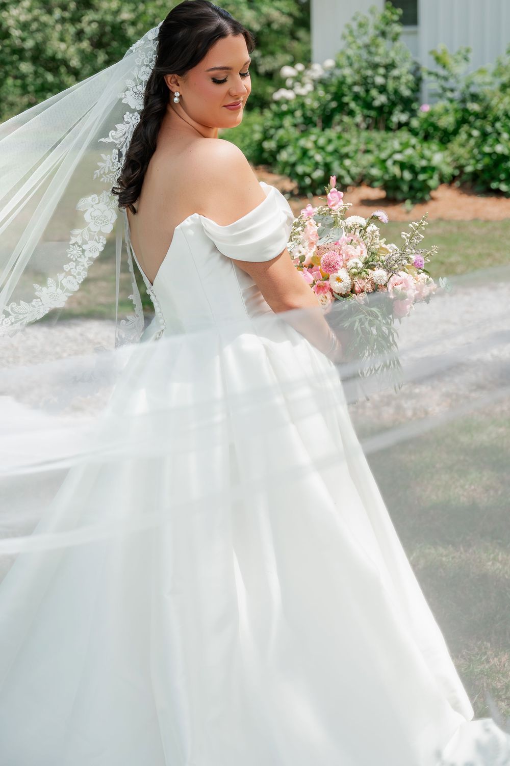 An elegant white wedding gown with off-shoulder details and lace veil flows in the breeze.