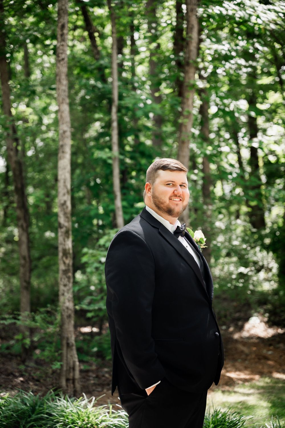 A groom in a classic black tuxedo poses in a wooded outdoor setting.