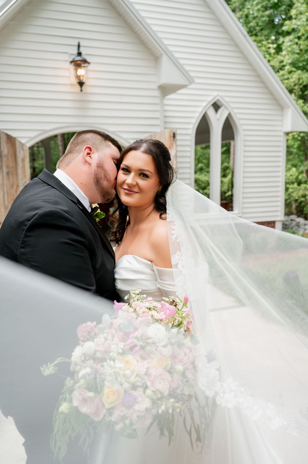 A bride in a flowing white dress holds a colorful bouquet while sharing an intimate moment with her groom.
