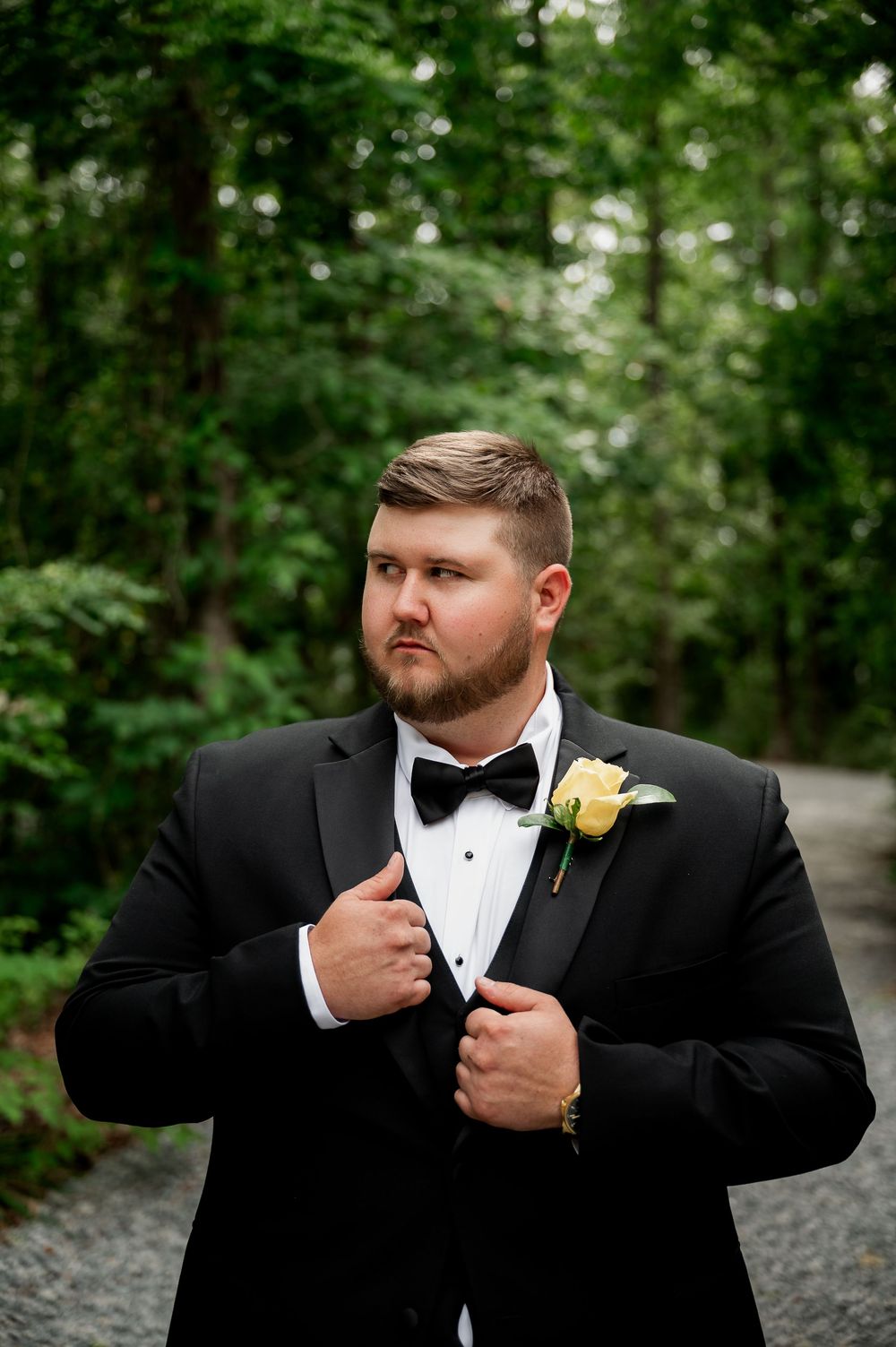 A groom in a black tuxedo and bow tie adjusts his collar during outdoor wedding photos.