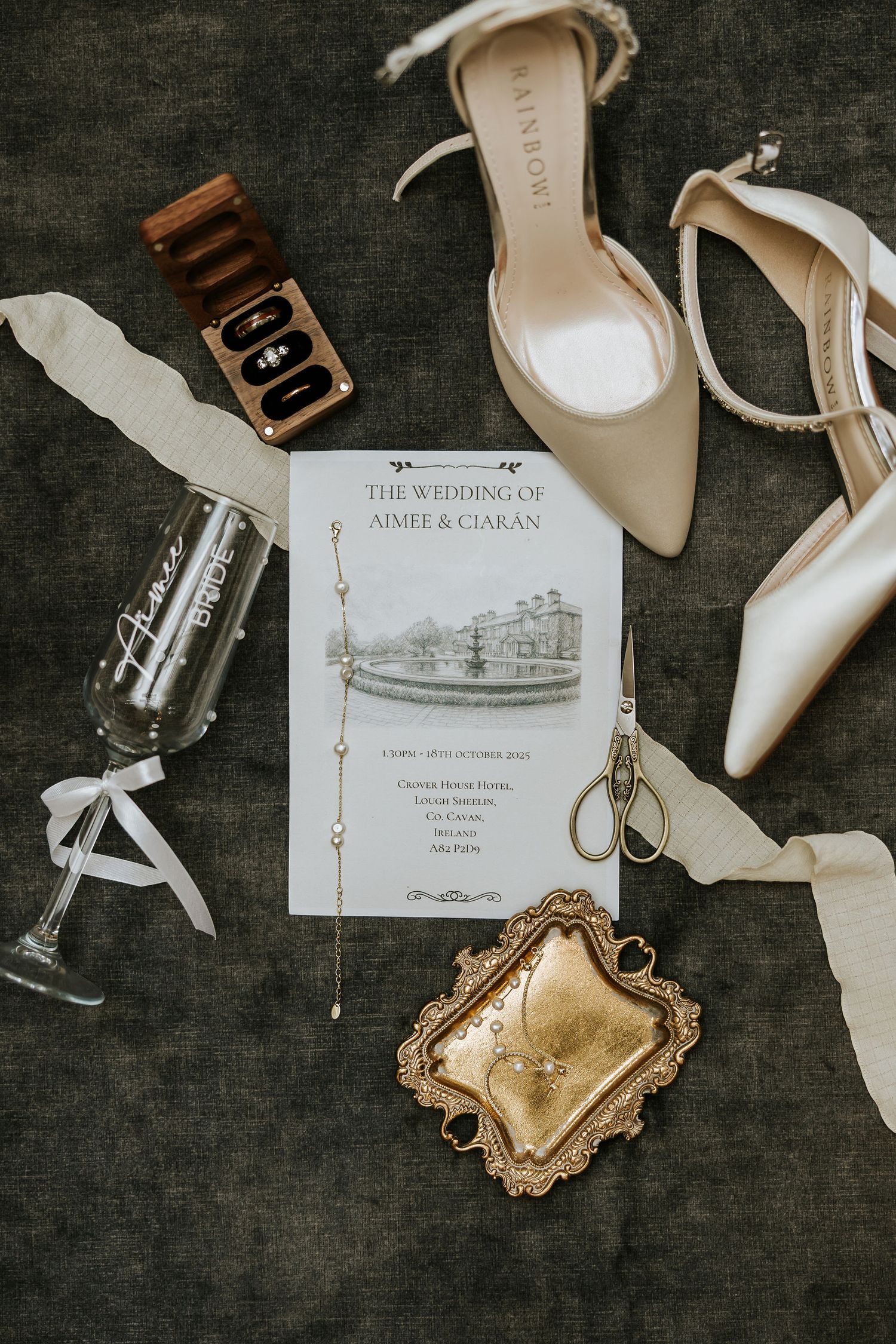Wedding details flatlay with cream heels, invitation, champagne glass and vintage accessories on dark background.