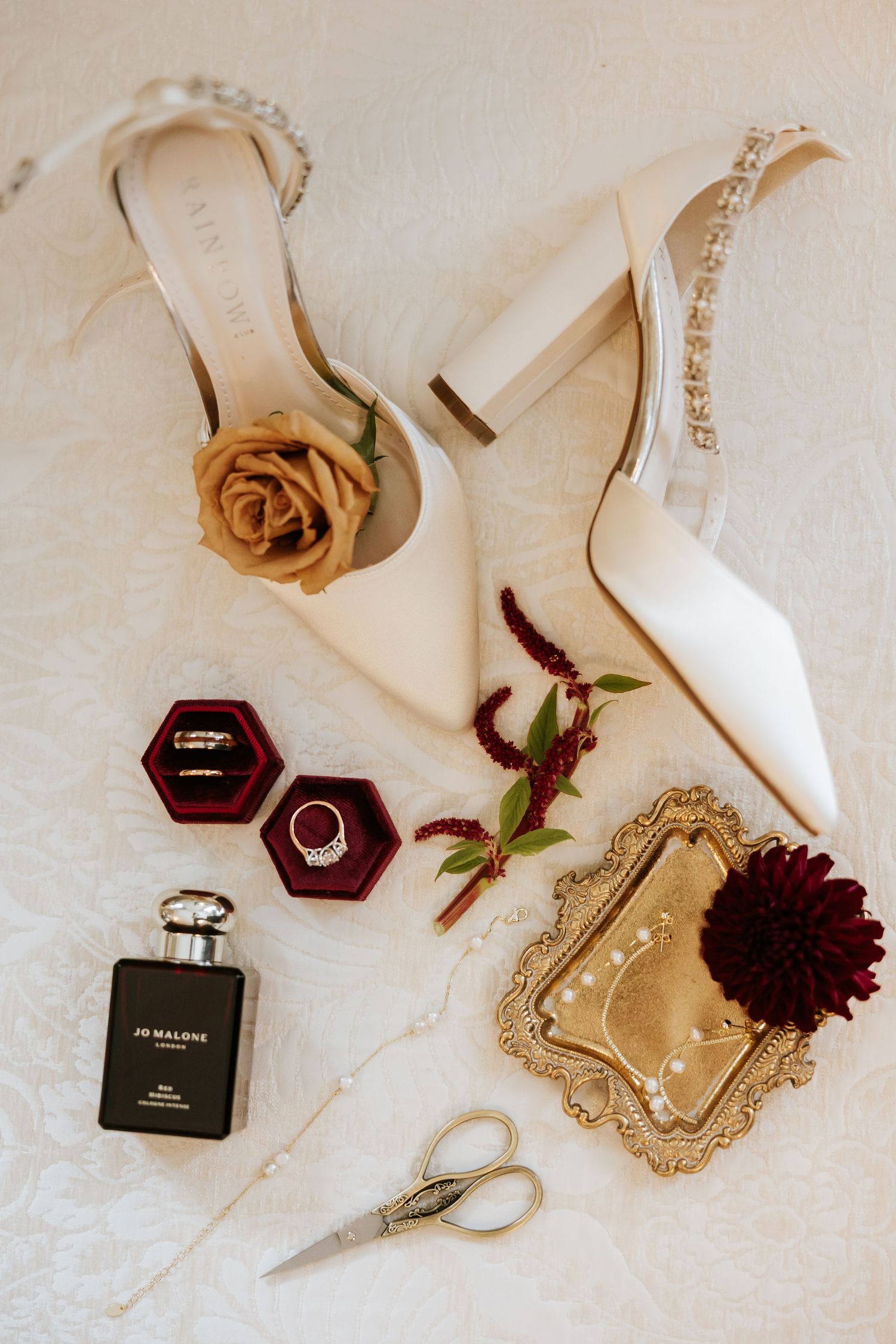 Flatlay arrangement of burgundy perfume bottle, white high heel shoes, and dark red flowers on beige background.