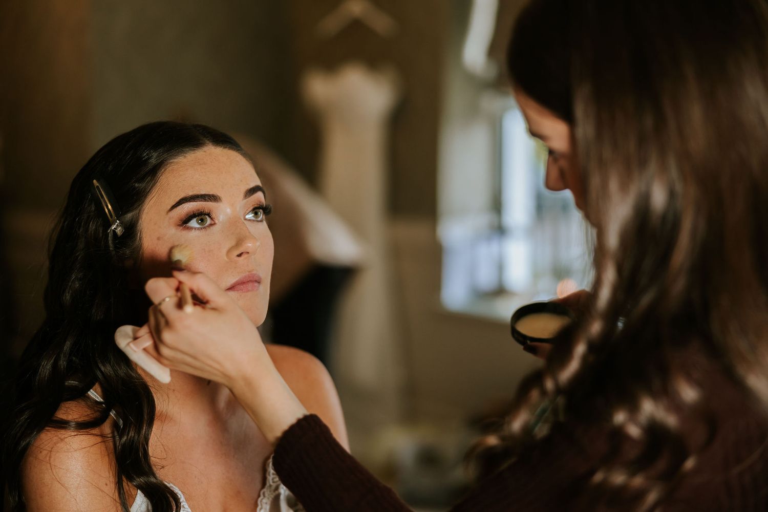 Professional makeup artist applies eyeshadow to client during bridal preparation in warm-lit room.