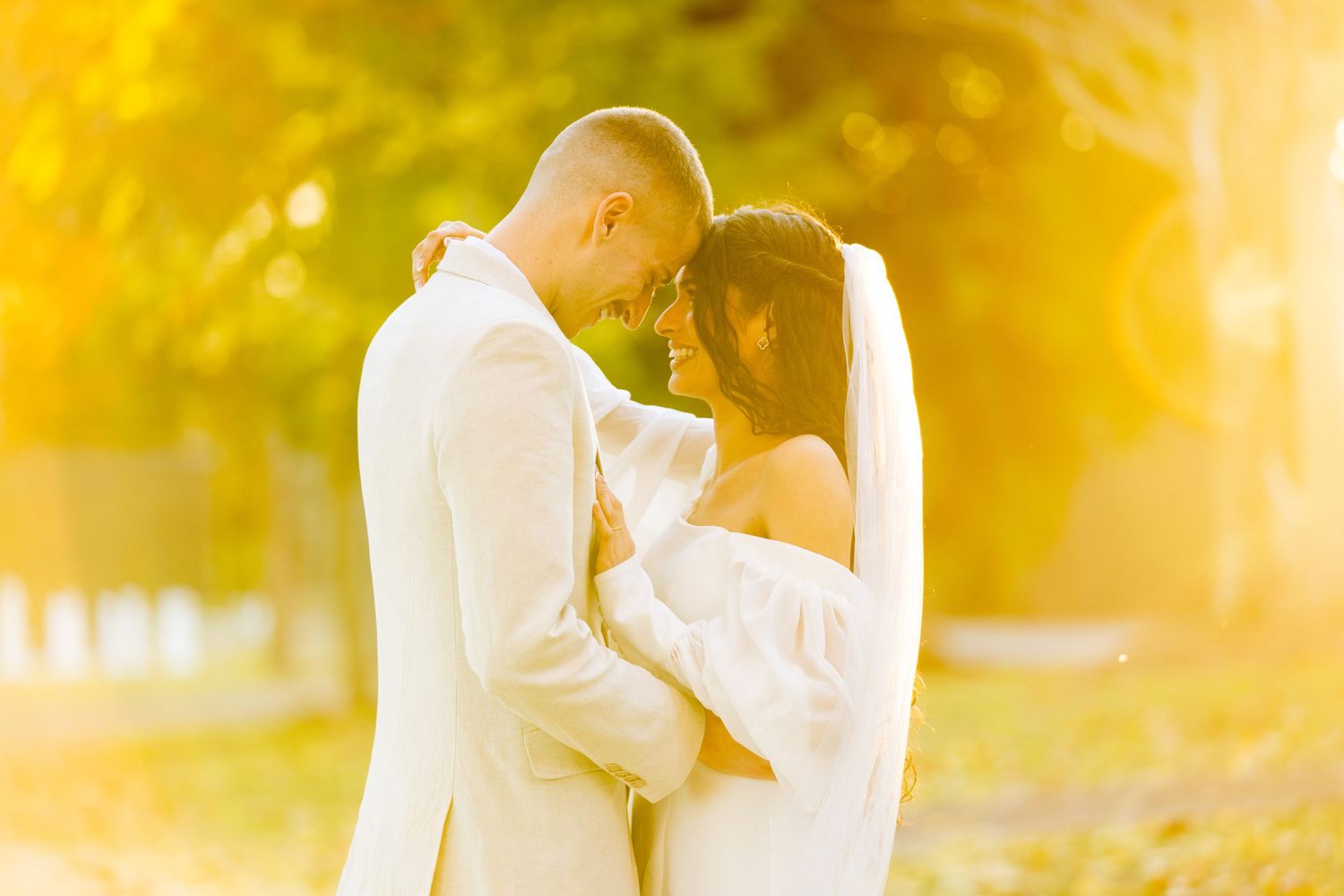 A romantic couple in white attire embraces outdoors during golden hour with soft sunlight creating a dreamy atmosphere.