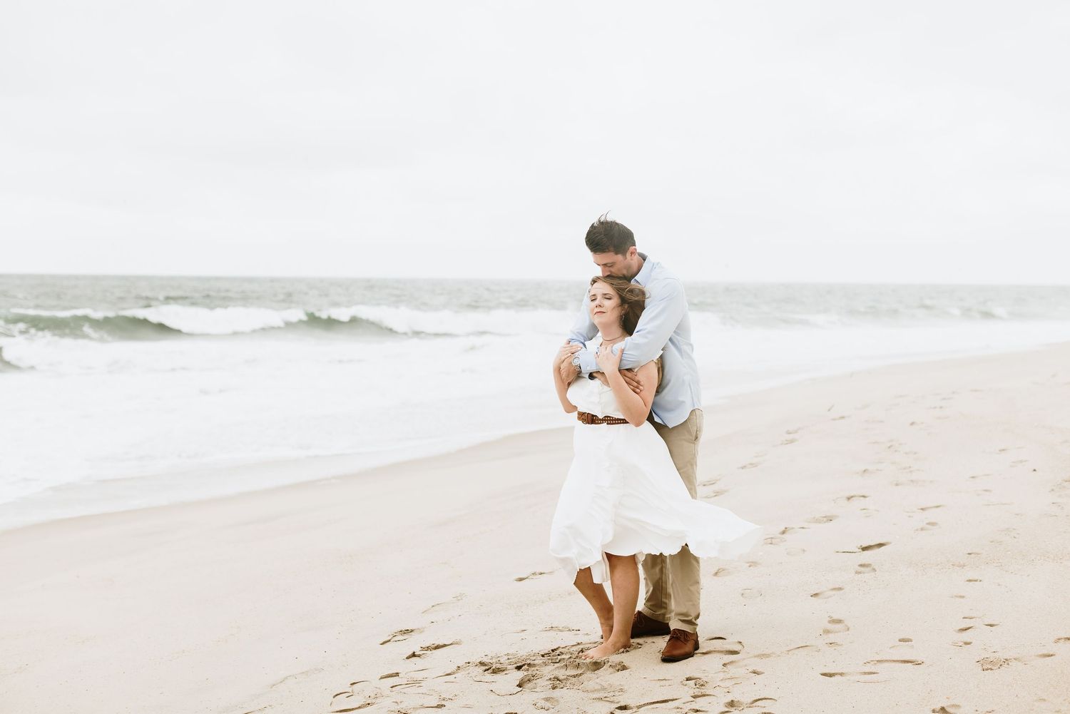 A man wearing a blue shirt and tan khakis embraces his fiancee from behind wearing a white dress for engagement photos in Point Pleasant, NJ