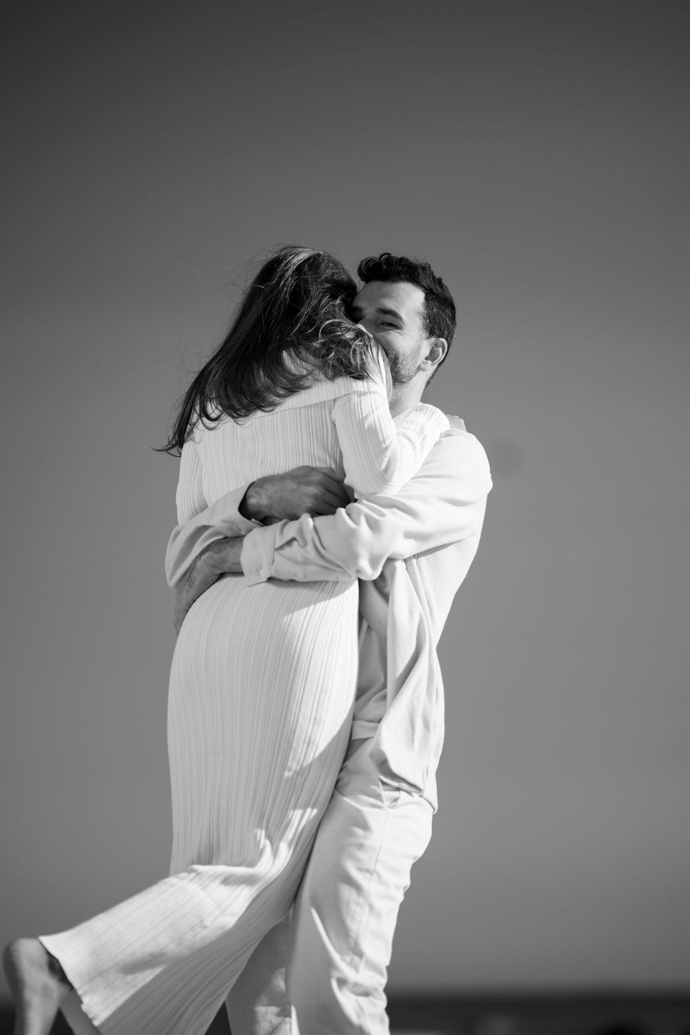 Black and white romantic photo of a couple kissing and embracing against a clear sky on a beach.