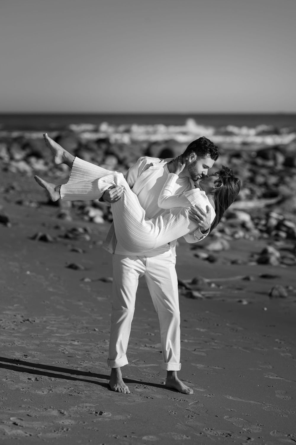 Black and white photo of a man lifting and kissing a woman on a sandy beach with rocks in background.
