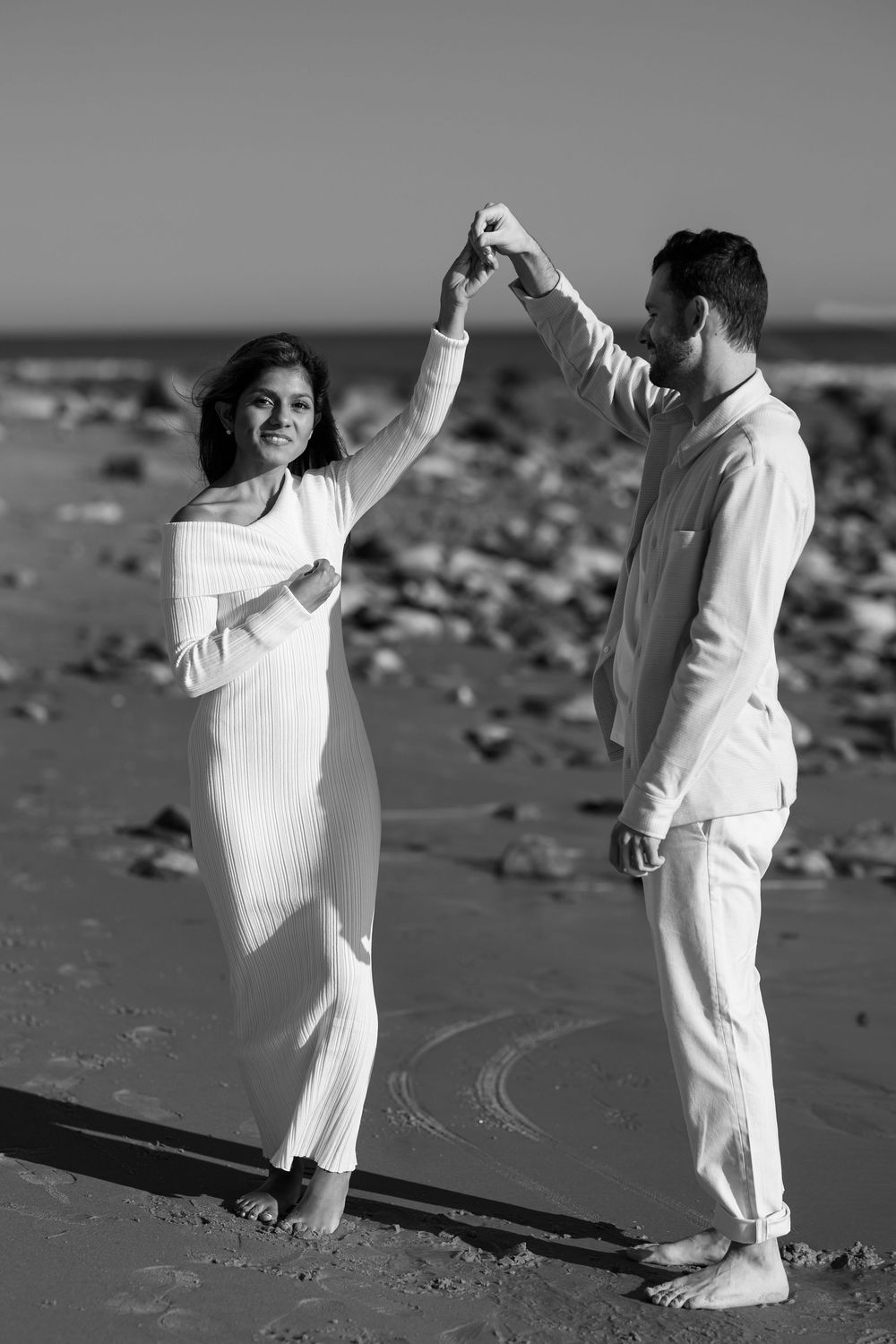 Black and white photo of a couple dancing on a beach, man spinning woman in a white dress by the ocean.