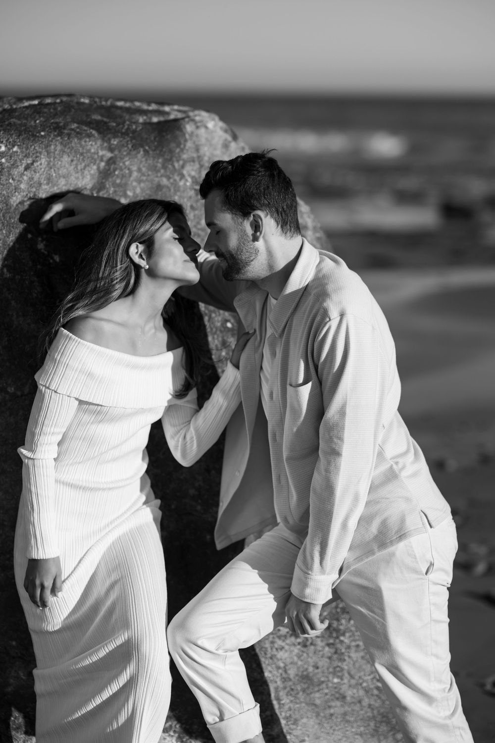 Black and white romantic couple gazing at each other leaning against a seaside boulder.