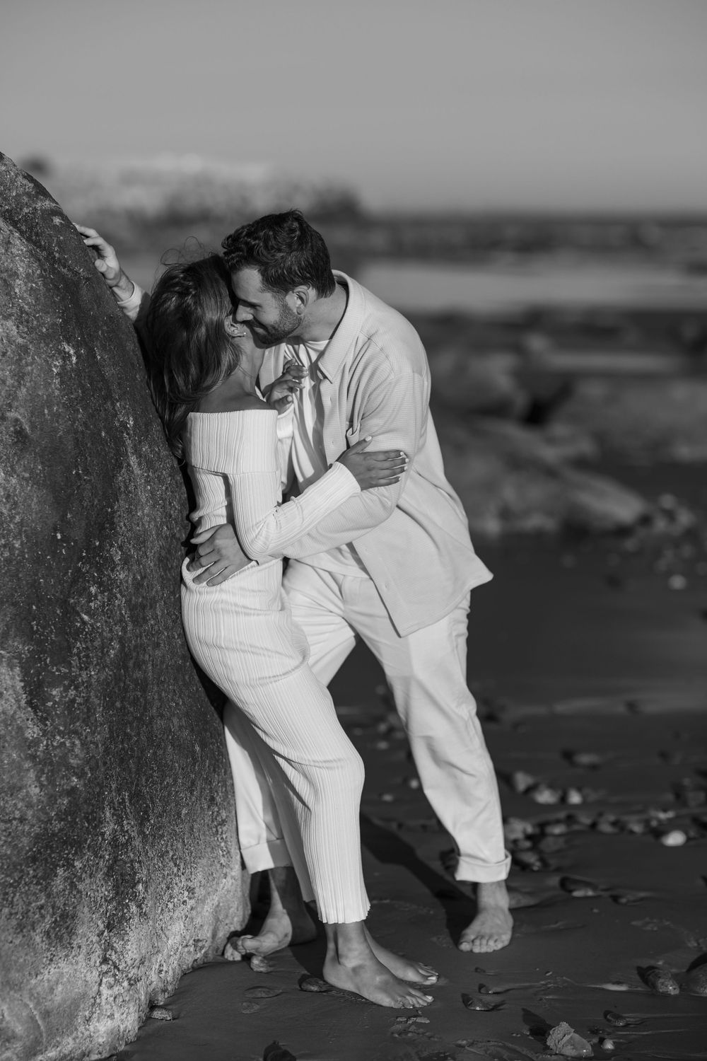 Black and white photo of a couple kissing against a large rock on a beach.