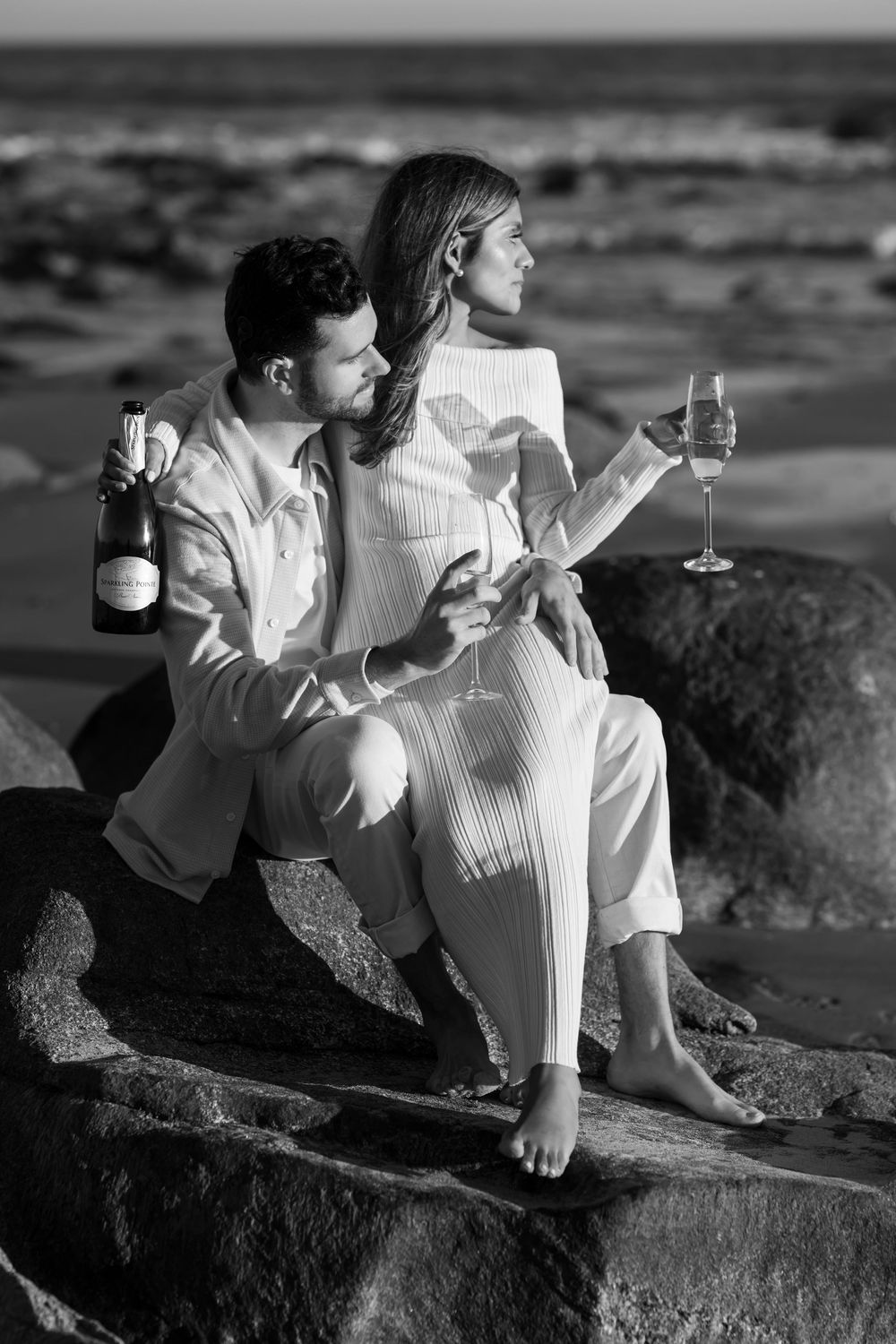 Black and white photo of a couple sitting on rocks by the ocean sharing champagne.