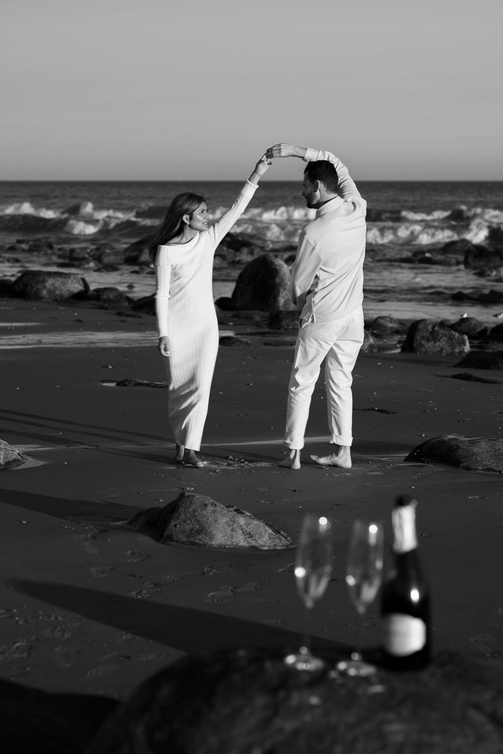 Black and white image of a couple dancing on the beach with champagne bottle in foreground.
