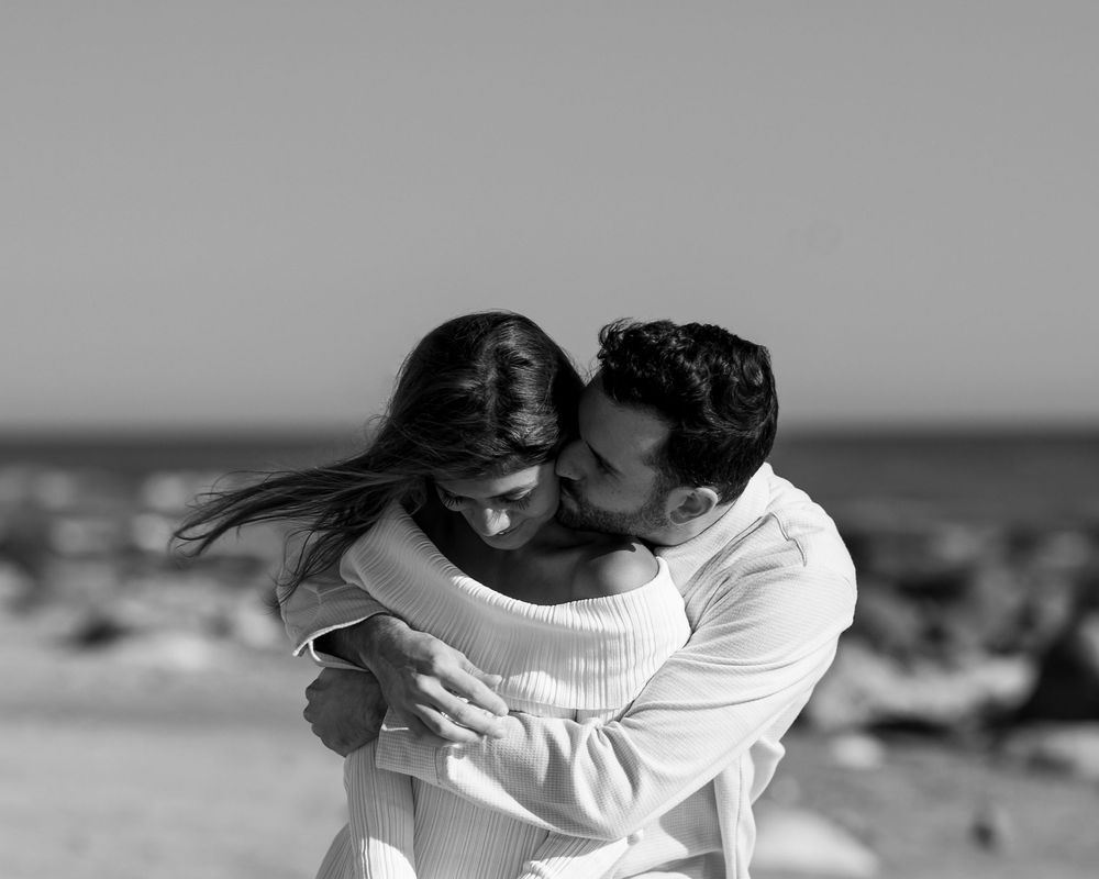 Black and white photo of a couple sharing a tender embrace on a sandy beach with ocean in background.