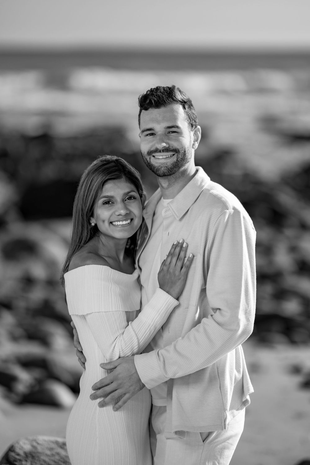 Black and white photo of a smiling couple embracing on a rocky beach, woman in white off-shoulder dress.