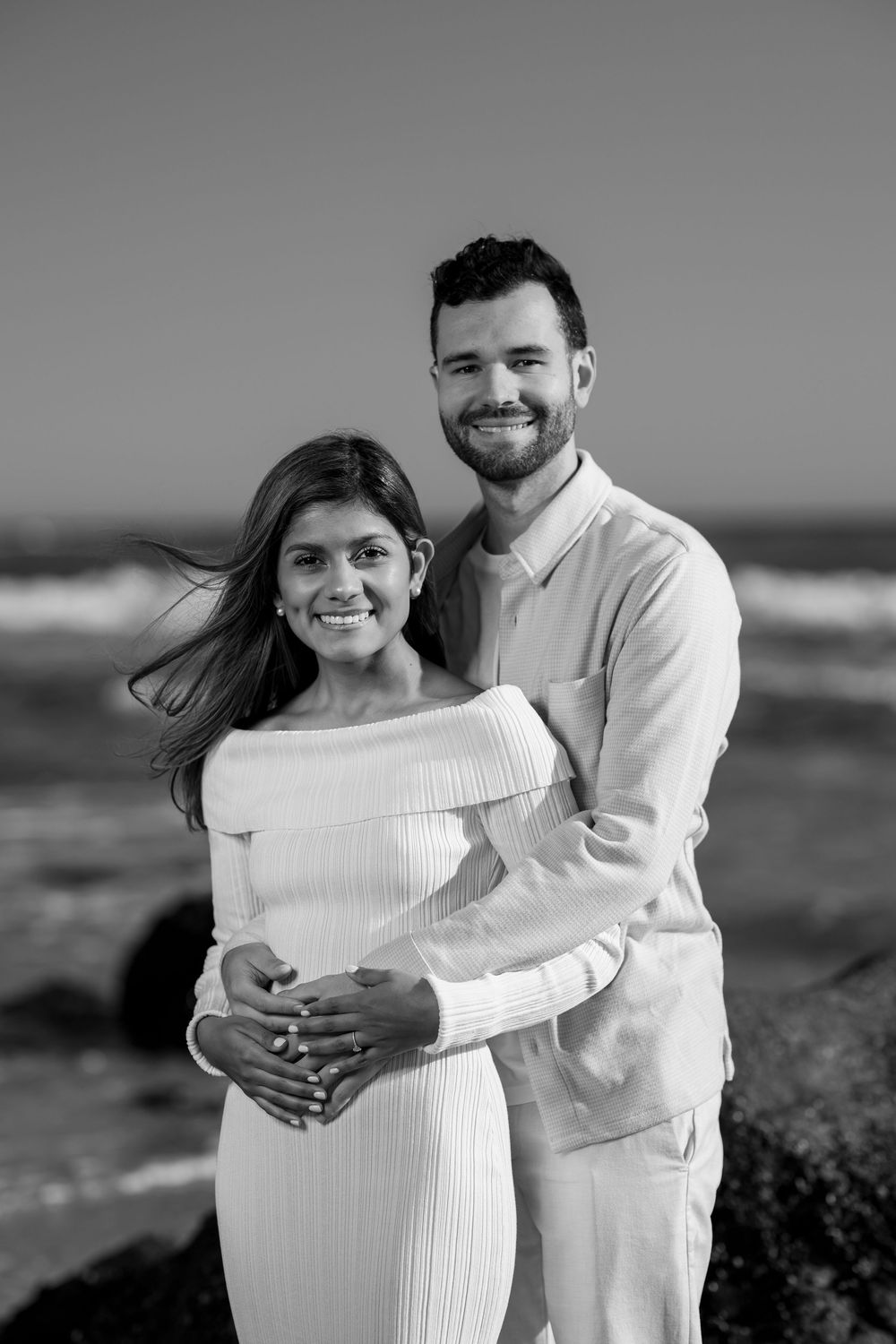 Black and white portrait of a happy couple embracing on a rocky beach with ocean waves behind them.