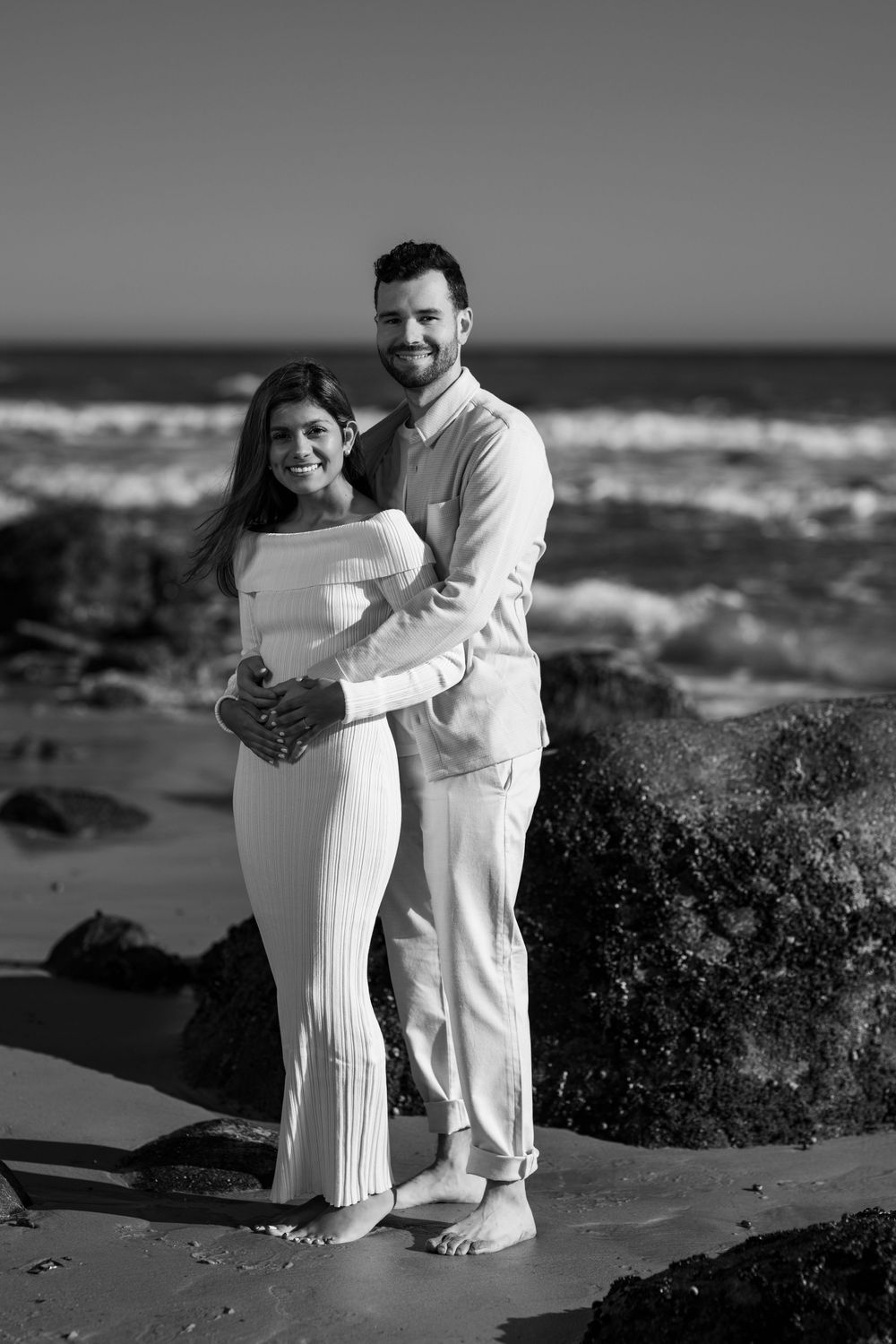 Black and white photo of a couple holding each other close on a sunny beach with rocky shoreline.