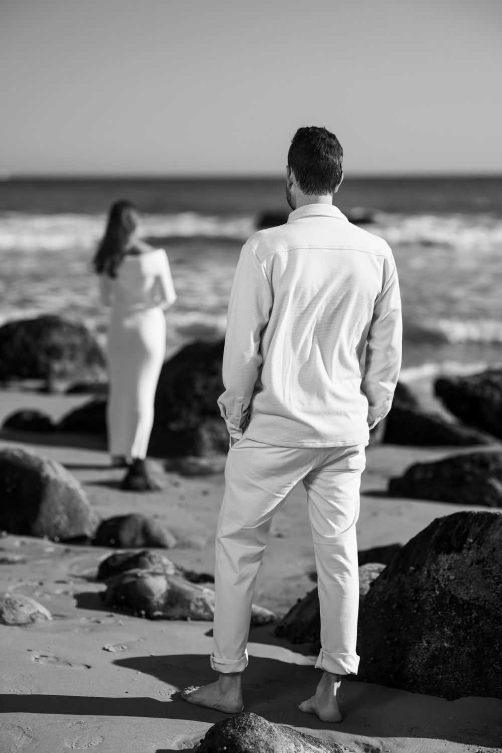 Black and white photo of a couple standing apart on a rocky beach, both dressed in white, facing the ocean.