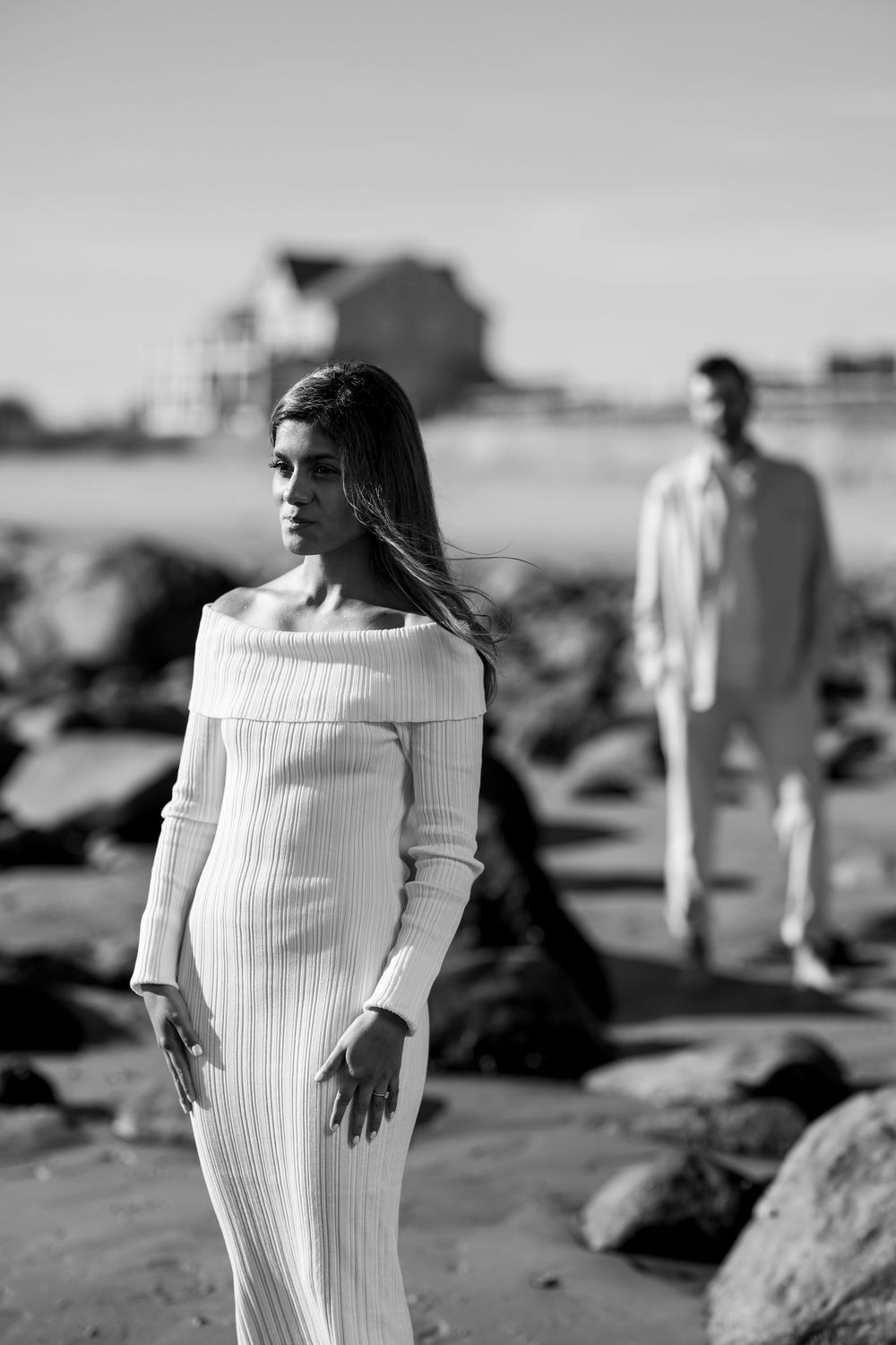 Black and white photo of a woman in a white ribbed dress posing on a beach with a man standing behind her.