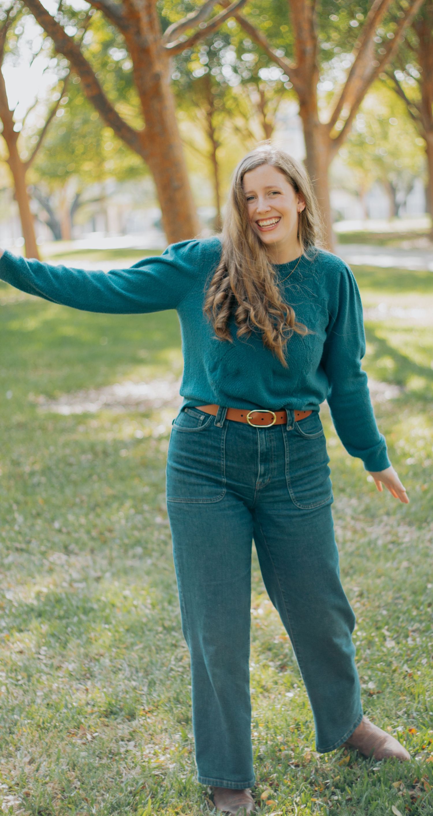 Someone wearing a teal sweater and jeans poses casually in a sunny park with autumn trees in the background.