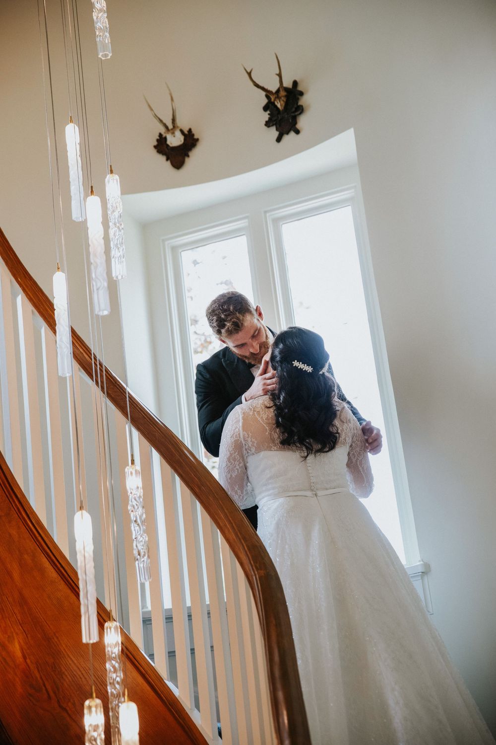 A romantic wedding moment captured on a staircase with elegant pendant lights and wooden railings.