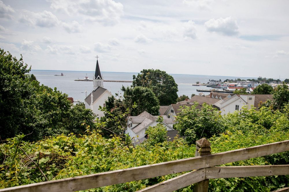 Panoramic coastal view showing multiple houses nestled along a hillside overlooking a harbor with boats and blue waters on Mackinac Island.