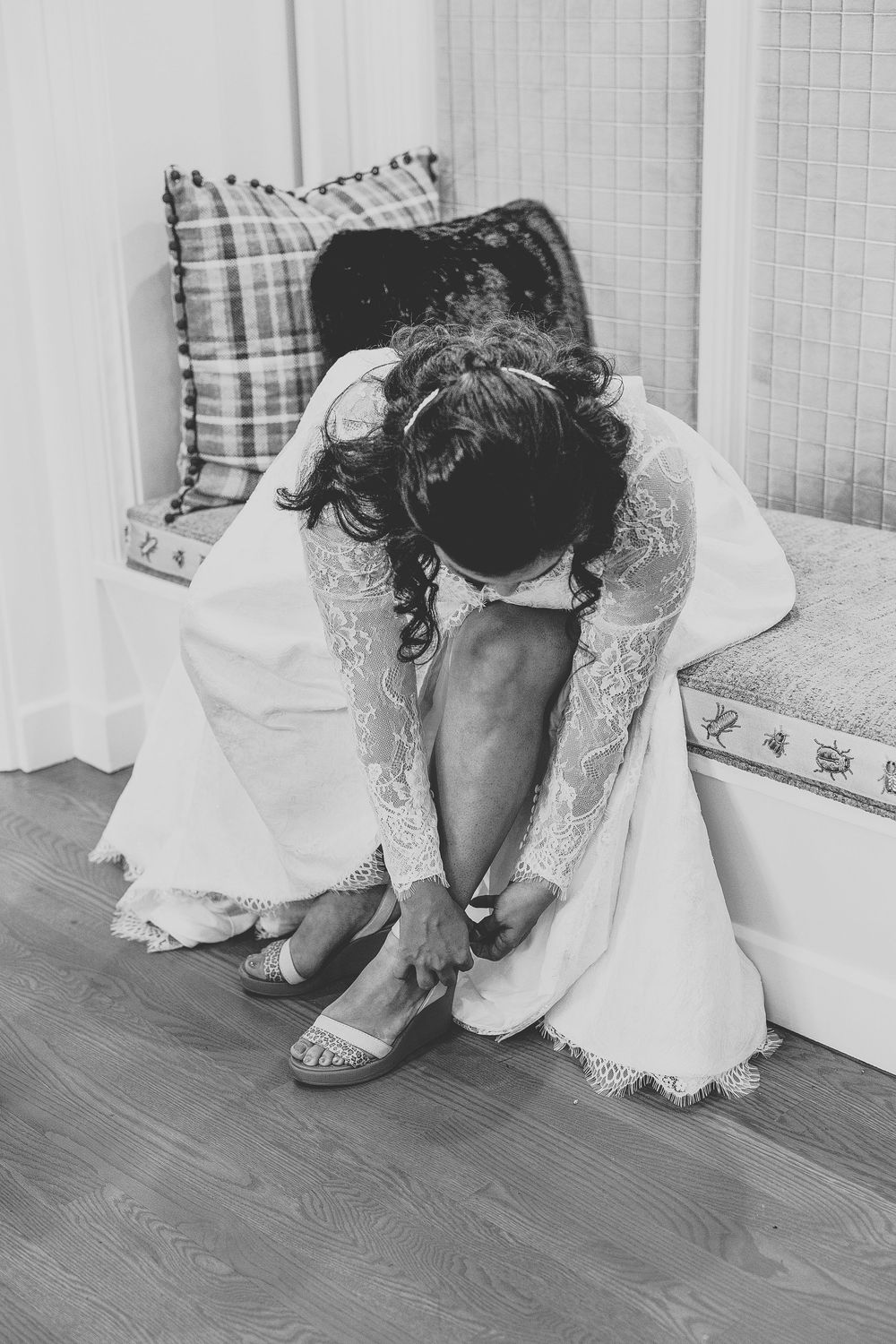 Black and white photo of a bride in a white lace dress fastening their high heal while seated on a bench.