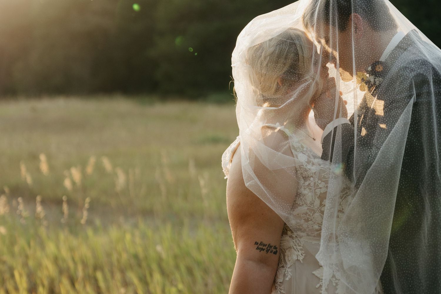 A romantic wedding couple embraces in a sun-drenched meadow during golden hour, with tall grass swaying in the breeze.
