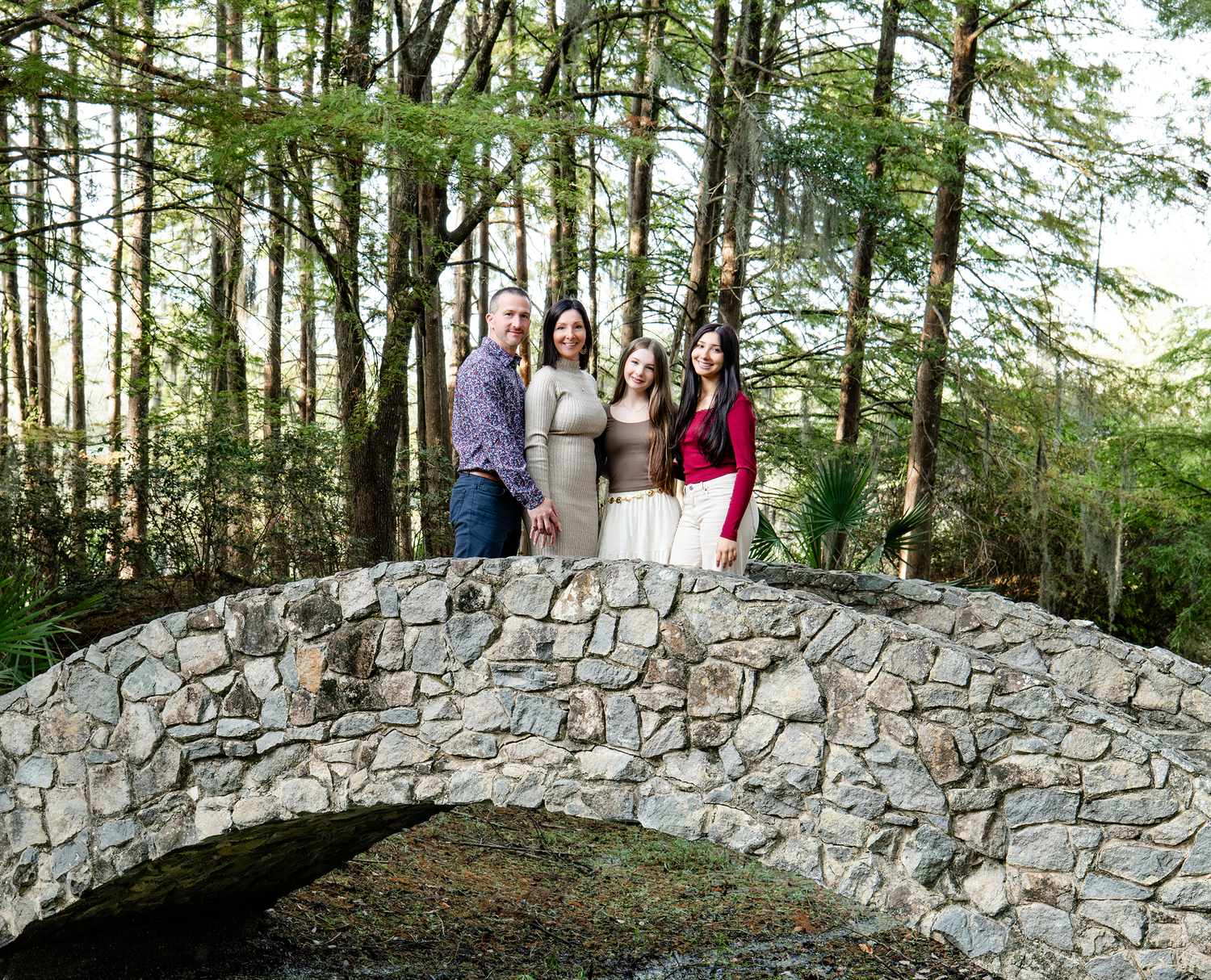 Family of four smiling together on a rustic stone arch bridge surrounded by tall green trees.