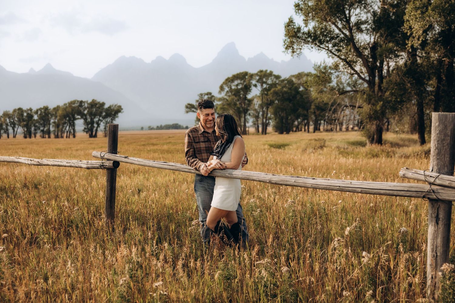 A couple embraces in a golden field during sunset with mountains visible in the background.