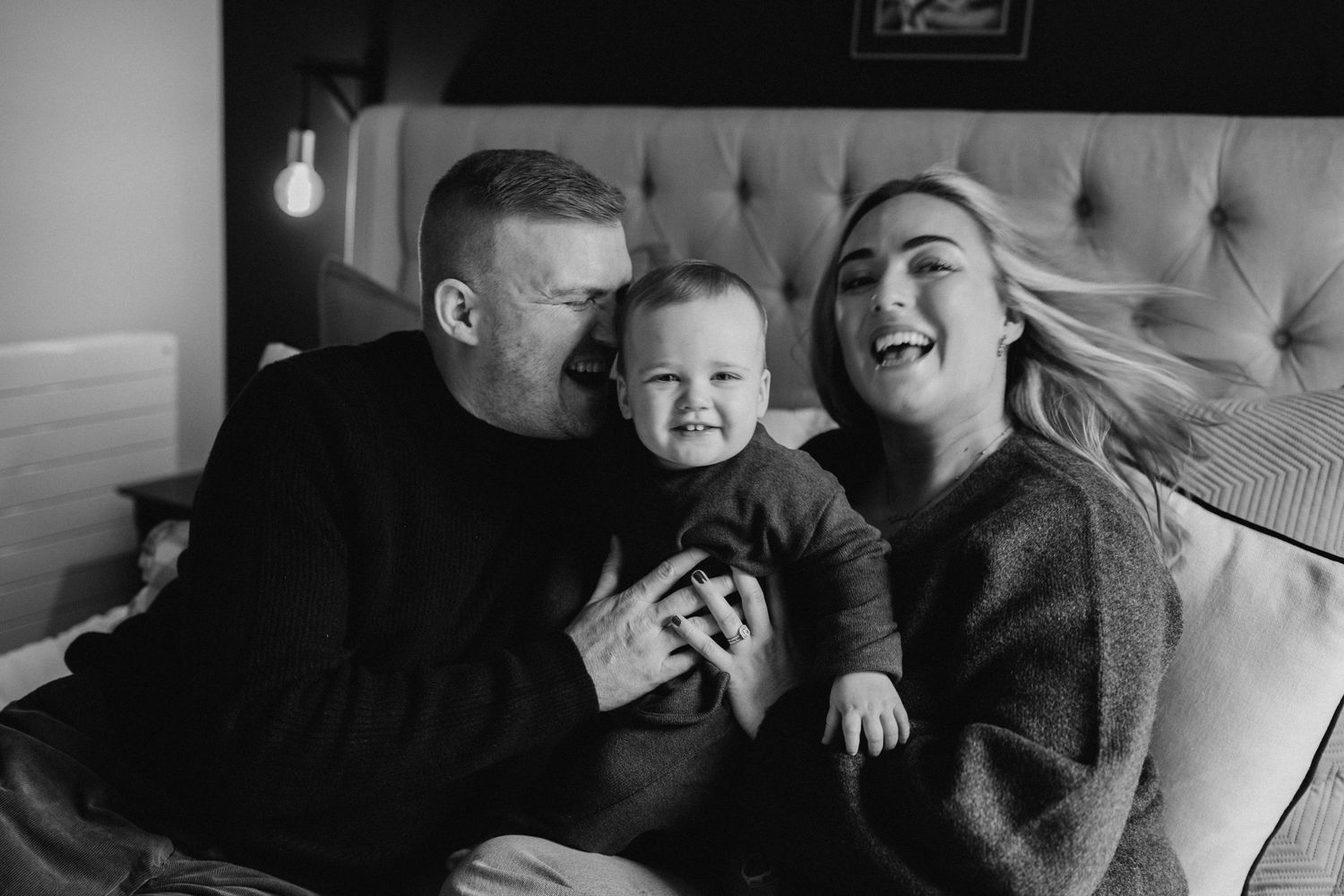 a family of three sitting on the bed, the baby an his mother a smiling in the camera while the father is kissing the baby
