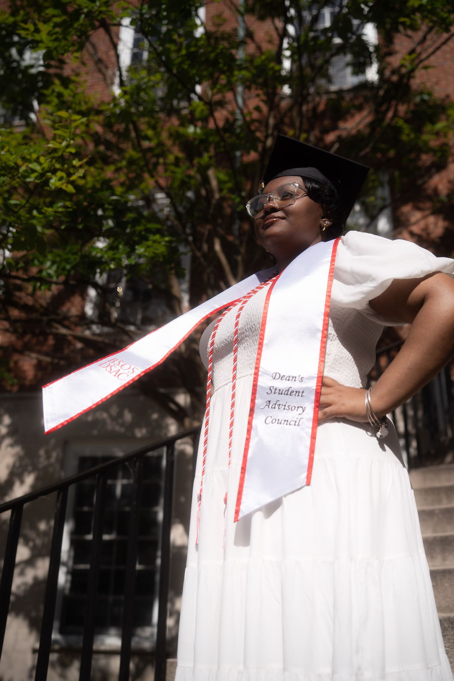Graduate in white dress and graduation stole poses triumphantly against brick building backdrop.