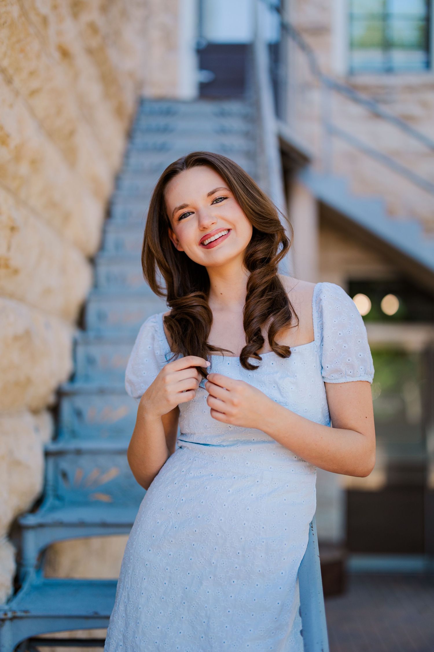 A young brunette woman in a light blue dress smiles near stone stairs in an outdoor urban setting.