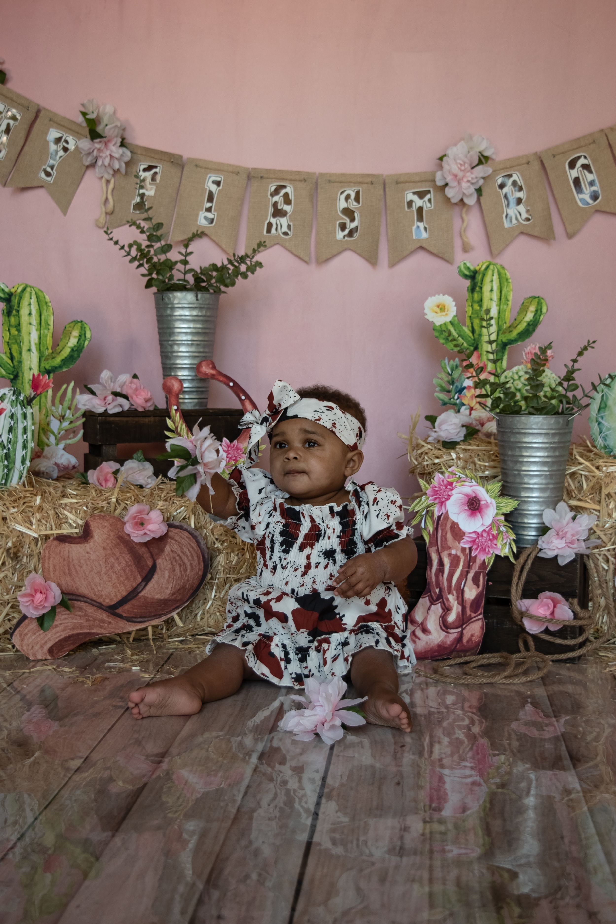 Rustic western-themed nursery setup with cowprint outfit, cactus decorations, pink flowers and burlap banner on pink wall.
