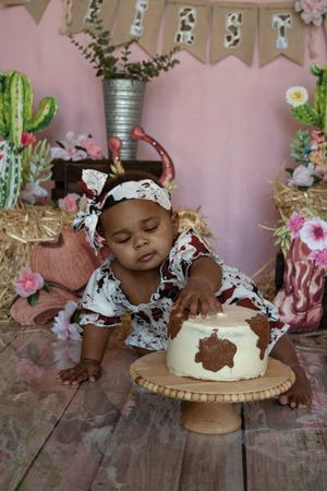 Baby in floral outfit reaching for cake on wooden stand with cactus and pink floral party decorations in background.