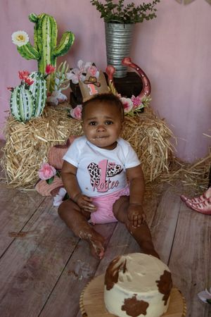 Western-themed baby photo session with cactus decorations, hay bales, and rustic wood flooring in pink tones.