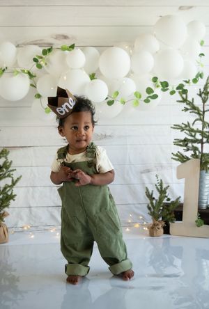 Small child in green overalls stands against white wall decorated with twinkle lights and white balloons.