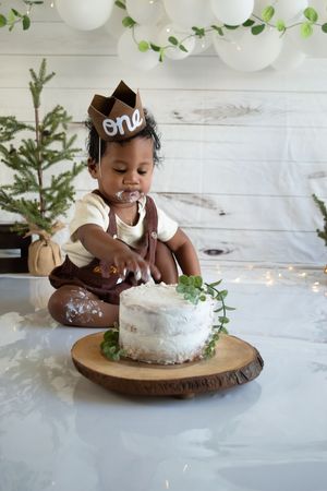 White frosted cake sits on wooden platter with greenery and white balloon decorations in background.