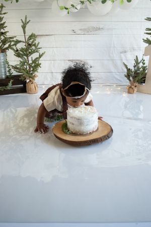 White frosted cake with greenery decorations sits on a wooden cake stand against a rustic white background.
