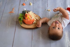 Baby lies on wooden floor while reaching for colorful food bowls during mealtime.