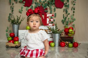 A small child in a white and red dress sits among roses and apples in a vintage-styled photoshoot setting.