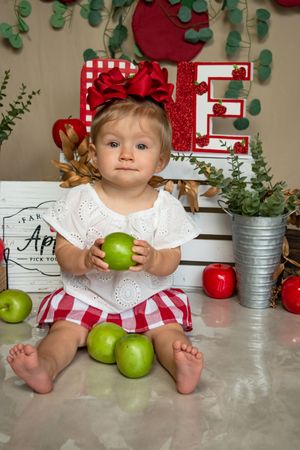 A baby in a white dress sits with green apples in a rustic kitchen setting with red and green decor.